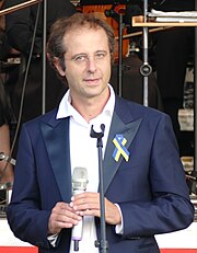 Luigi Gaggero holding a microphone and wearing a bow in the Ukrainian national colours on his jacket.