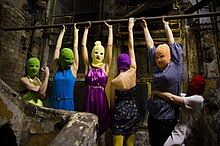 A group of 6 women wearing ski masks and multicolored clothes in a small, grubby brick space. Four of them hang by their arms from a bar, one flexes her muscles, and another grabs one of the hanging women around the waist.