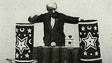 Georges Méliès stands behind a table, on which are placed various lavishly decorated props for a magic show.