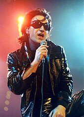 Bono with black hair, black sunglasses, and black leather attire speaks into a microphone.