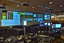 A dark office filled with computers; screens of financial data fill one wall