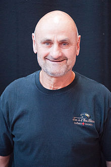 Photographie couleur d’un homme, de tronc, chauve, souriant, avec une barbe de 3 jours blanche et un t-shirt bleu marine