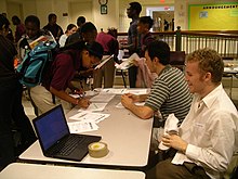 Two young white men sit at a table with several teenage African American students mingle around it, and one signs a paper on it. Also on the table is a laptop.