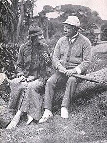 A woman with braided hair and a headband, dressed in a long skirt and dark blouse, sitting on a large log with an older man wearing a hat