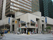 A block-shaped concrete building sits on a city street corner in front of a steel-and-glass skyscraper