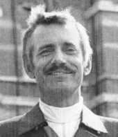 Photo en noir et blanc d'un homme moustachu d'une quarantaine d'années, souriant, vêtu d'un col roulé et d'une veste à col pelle à tarte.