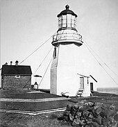 photo noir et blanc avec le phare, avec ses étais, au centre de l'image, sur la gauche une maison, au premier plan un muret en pierre sur la droite et la toiture de la citerne d'eau à gauche. En arrière-plan c'est majoritairement le ciel avec une petite vue sur la mer à droite