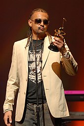 A picture of a man wearing a beige jacket and holding a silver award statue