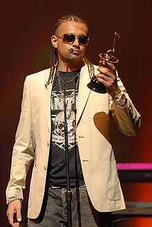 A young black man in a black T-shirt, white jacket and black trousers, receiving an award standing in front of a microphone.