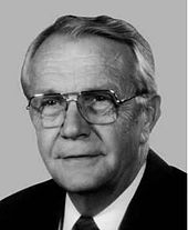 A man with receding, gray hair and glasses wearing a white shirt with a black tie and jacket