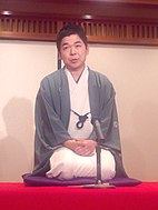 A Japanese man kneeling wearing a cream kimono and a blue jacket