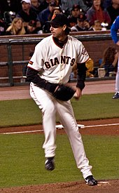 A man in a white baseball uniform with the word "GIANTS" written across it prepares to throw a baseball with his left hand to home plate during a game.