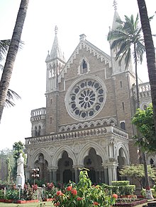 The front of a tall neo-Gothic building with a porch is seen behind palm trees.