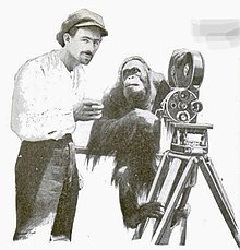 Man with oversize painter's cap and a mustache holding a cup of coffee; orangutan hanging off a film camera mounted on a tripod