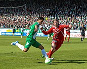 A soccer player dressed in green and white runs with the ball. A player in red and white tries to take the ball. In the background there are stands filled with Hammarby fans.