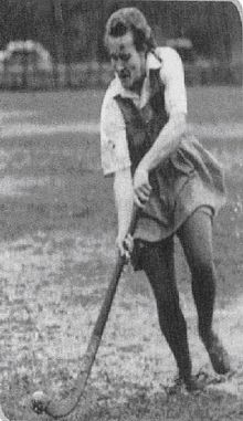 black-and-white photo of a female hockey player wearing a knee-length skirt and elbow length shirt. She is wearing dark colour tights. She is standing upright while holding the hockey stick pointed down at the ground. She is in a running position and about to hit a ball that is shown in the lower left hand corner.