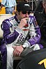 A racing driver in a purple-and-white firesuit, black cap and dark glasses broods while waiting in the pits.