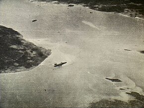 Black and white aerial photograph showing a body of water with a large warship near the shore