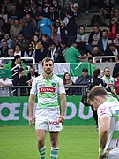 Photo en couleur montrant un joueur de rugby habillé en blanc et vert marchant sur une pelouse, au fond des spectateurs.