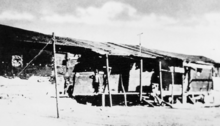 A black and white photo from around 1907, depicting 2 people sitting on the porch of a crude shanty.