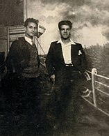 Photographie en noir et blanc de deux jeunes hommes debout, vêtus de costumes et regardant l'objectif.