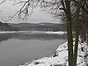 A grey lake under grey skies with snow and ice blocks on its shore and snow covered low mountains in the background