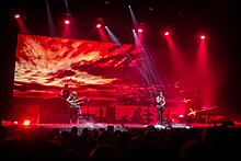 Trois membres d'un groupe de rock jouant devant un écran montrant un coucher de soleil.