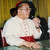 Jaime Cardinal Sin of the Philippines shown wearing a white cassock with a red belt and skullcap holding a microphone