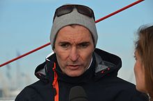 Portrait de face, veste de quart noire, bonnet gris, répondant à une journaliste.