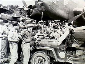 Approximately 15 men wearing military uniforms in discussion around a jeep, parked in front of a twin-engined aircraft