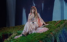 Taylor Swift singing while sitting on moss, dressed in a floating dress