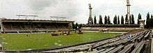 Photographie d'un stade de football vu d'un angle des tribunes, des engins de chantier sont au centre du terrain.