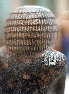 Close-up of the back of the head and shoulder of a granite statue showing white inscribed hieroglyphs on a reddiwh-brown background