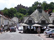 A small single story building with a pyramid shaped roof, to the side of a road lined with buildings. Some private small cars visible. Trees in the distance with the skyline of Dunster Castle.