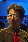 A 50-year-old Japanese man with black-brown hair in a brown suit with a red tie, smiling away from the camera.