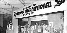 Black and white photo of a banner that says "Welcome to Flagship International Sports TV" with a line of people in the background and a man wearing a war bonnet in the foreground