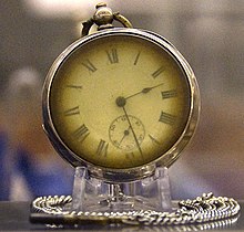 Photograph of a brass pocket watch on a stand, with a silver chain curled around the base. The watch's hands read 2:28.