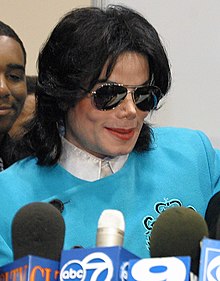 Jackson is wearing a white shirt and a light blue pullover. His skin is light and his nose is thin. He's wearing make-up and sun glasses. His hair is straight. Some microphones are standing on a lectern in front of him.