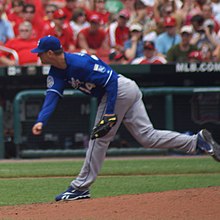 A man in a blue baseball jersey pitches a baseball to home plate with his right hand.