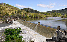 A large river flows over a dam and into a churning pool below. Green hills line the far shore.