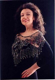 A photograph of a smiling white woman with long dark wavy hair, posing in an elegant black evening dress