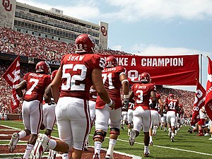 Football players run onto a football field in two rows under a crimson banner.