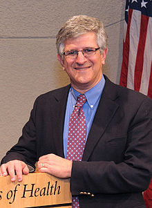 grey-haired white male leaning on podium and smiling at camera