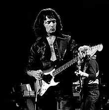 A guitarist, Ritchie Blackmore, is shown playing a Fender electric guitar onstage. He has long hair.