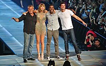 Four people—a middle aged white man in a striped blackg shirt and jeans, a young white woman in heels and a sparkling dress, a black man in a grey shirt and pants, and a white man in a white tee and jeans, posing together onstage