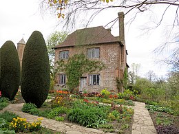 a cottage built of pink brick in the middle of a garden. Spring flowers and two tall Cyprus trees stand in front of the cottage