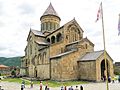 cathédrale en pierres de taille roses et sable à étages, surplombée par un clocher coiffé d'un toit de teinte mauve surmonté d'une croix orthodoxe. À l'arrière-plan, des montagnes boisées et à l'avant-plan à droite deux mâts ornés du drapeau géorgien (croix rouge sur fond blanc).