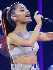 A woman with brown hair singing on stage wearing a lilac crop top and skirt