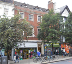 City street scene showing continuous development with a red brick, three storey building having two modern shop fronts at street level and a doorway in between