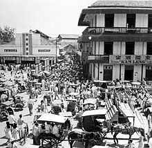 Crowd at a busy street intersection. There are horse-drawn carriages in the foreground while a three-story building (with the sign "Kam Leng") and a single story building (with the sign "Chunghua Bioscoop") stand in the background on adjacent corners of the intersection.